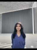 A young indian woman with long dark brown hair wearing a v-neck blue sweater stands with her hands behind her back in front of a gray building and smiles at the camera.